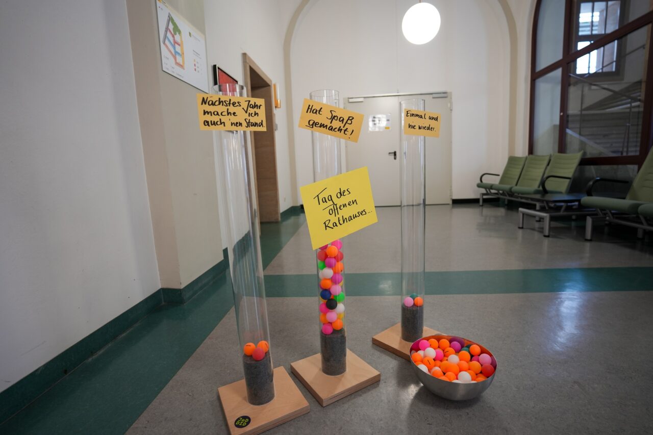 KI generiert: Das Bild zeigt drei durchsichtige Röhren, die mit bunten Bällen gefüllt sind, und daneben eine Schale mit weiteren Bällen. Die Röhren sind mit Schildern versehen, die Aussagen über den "Tag des offenen Rathauses" machen.